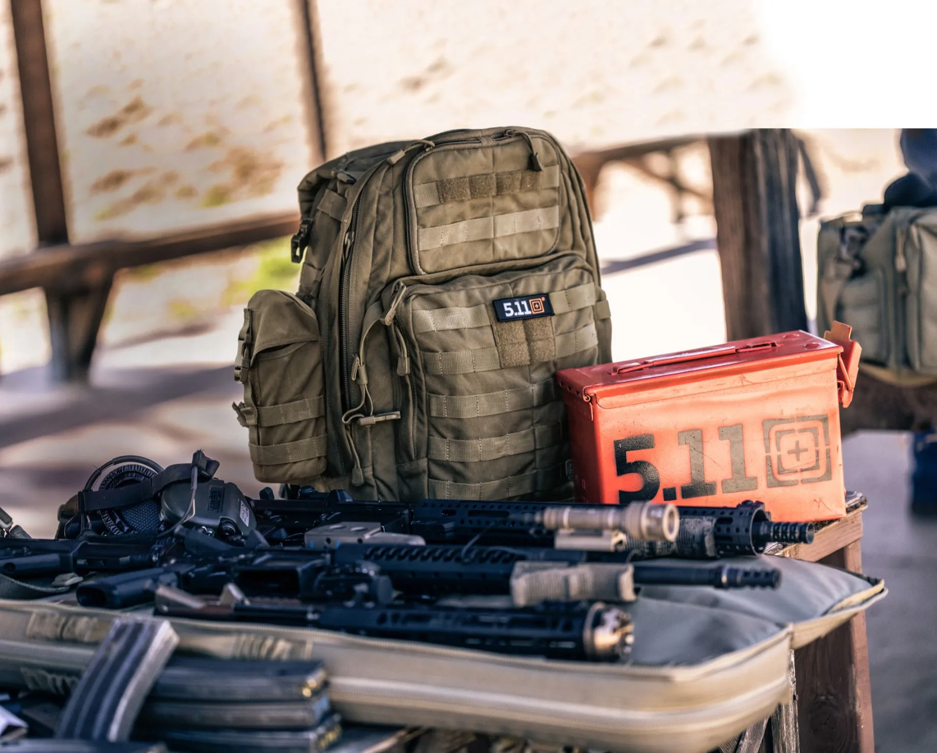 Tan 5.11 backpack and an orange 5.11 ammo can on a shooting range table with rifles in the foreground
