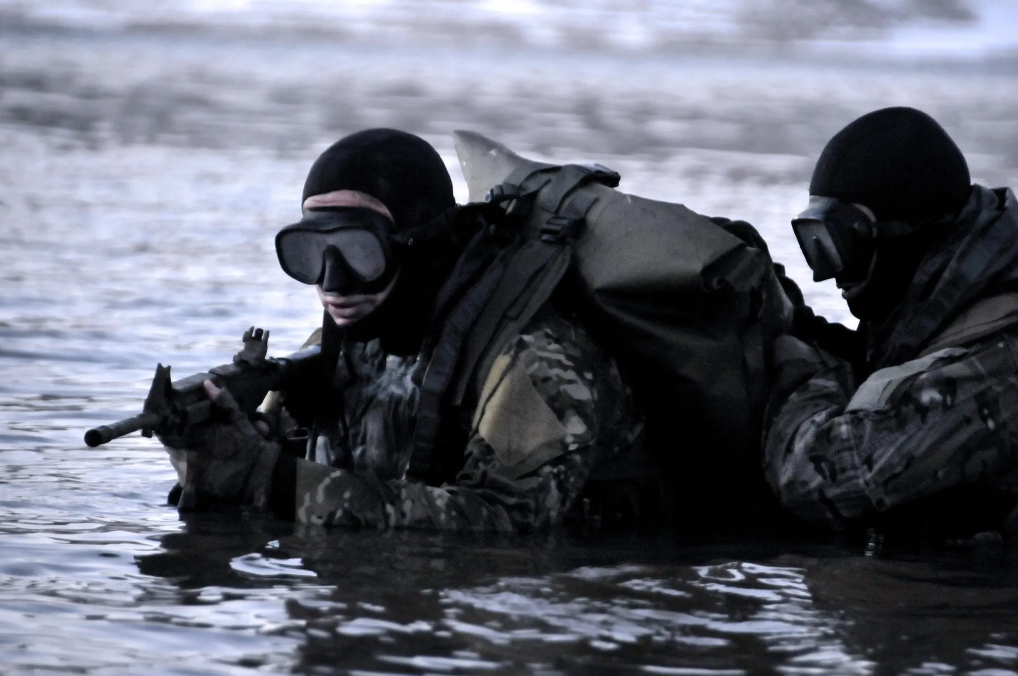 Two combat swimmers with masks and a waterproof Watershed bag wading through water with a rifle